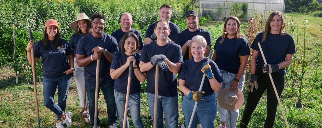 Several people, wearing dark t-shirts and jeans, holding shovels in a field on a sunny day.